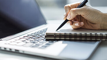 Ein Schreibblock liegt auf einem Laptop und eine schreibende Hand mit Stift 