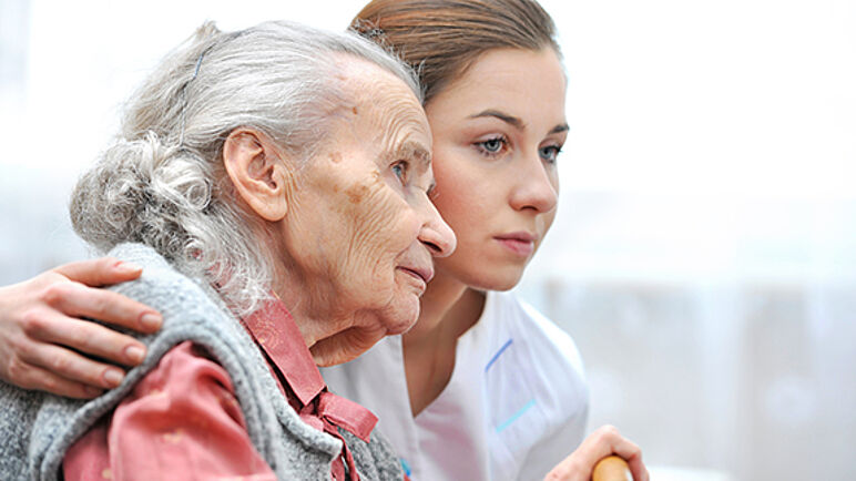 Eine Pflegerin umarmt die Seniorin und beide schauen nachdenklich in die Ferne
