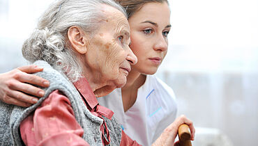 Eine Pflegerin umarmt die Seniorin und beide schauen nachdenklich in die Ferne