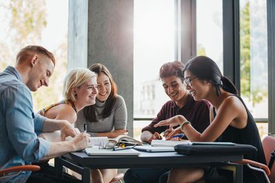 Eine Gruppe von Lernenden sitzt lachend zusammen an einem Tisch mit Büchern, Zetteln und Schreibutensilien