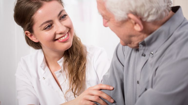 Eine Pflegerin spricht mit einem Senior, ihre Hand ist auf seinem Arm