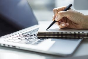 Ein Schreibblock liegt auf einem Laptop und eine schreibende Hand mit Stift 