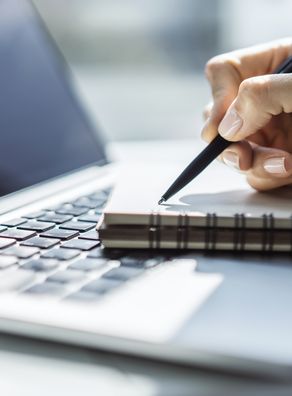 Ein Schreibblock liegt auf einem Laptop und eine schreibende Hand mit Stift 