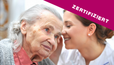 Eine Pflegerin flüstert der Seniorin etwas ins Ohr – im Blickfang steht "zertifiziert"
