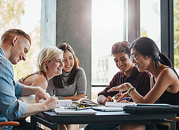 Eine Gruppe von Lernenden sitzt lachend zusammen an einem Tisch mit Büchern, Zetteln und Schreibutensilien