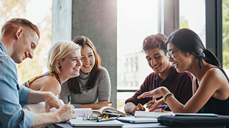 Eine Gruppe von Lernenden sitzt lachend zusammen an einem Tisch mit Büchern, Zetteln und Schreibutensilien